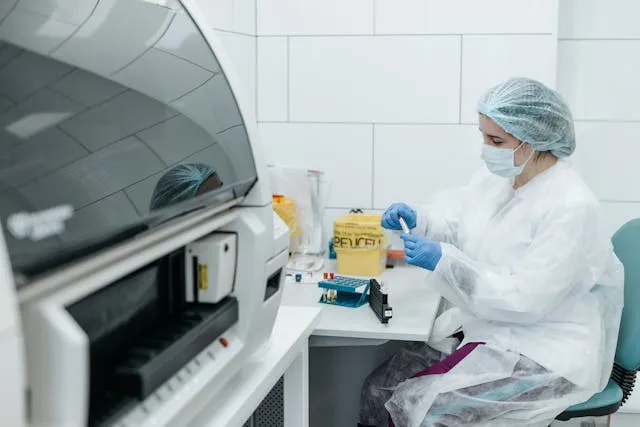 Lab technician wearing protective gear and gloves handling samples in a laboratory with testing equipment.