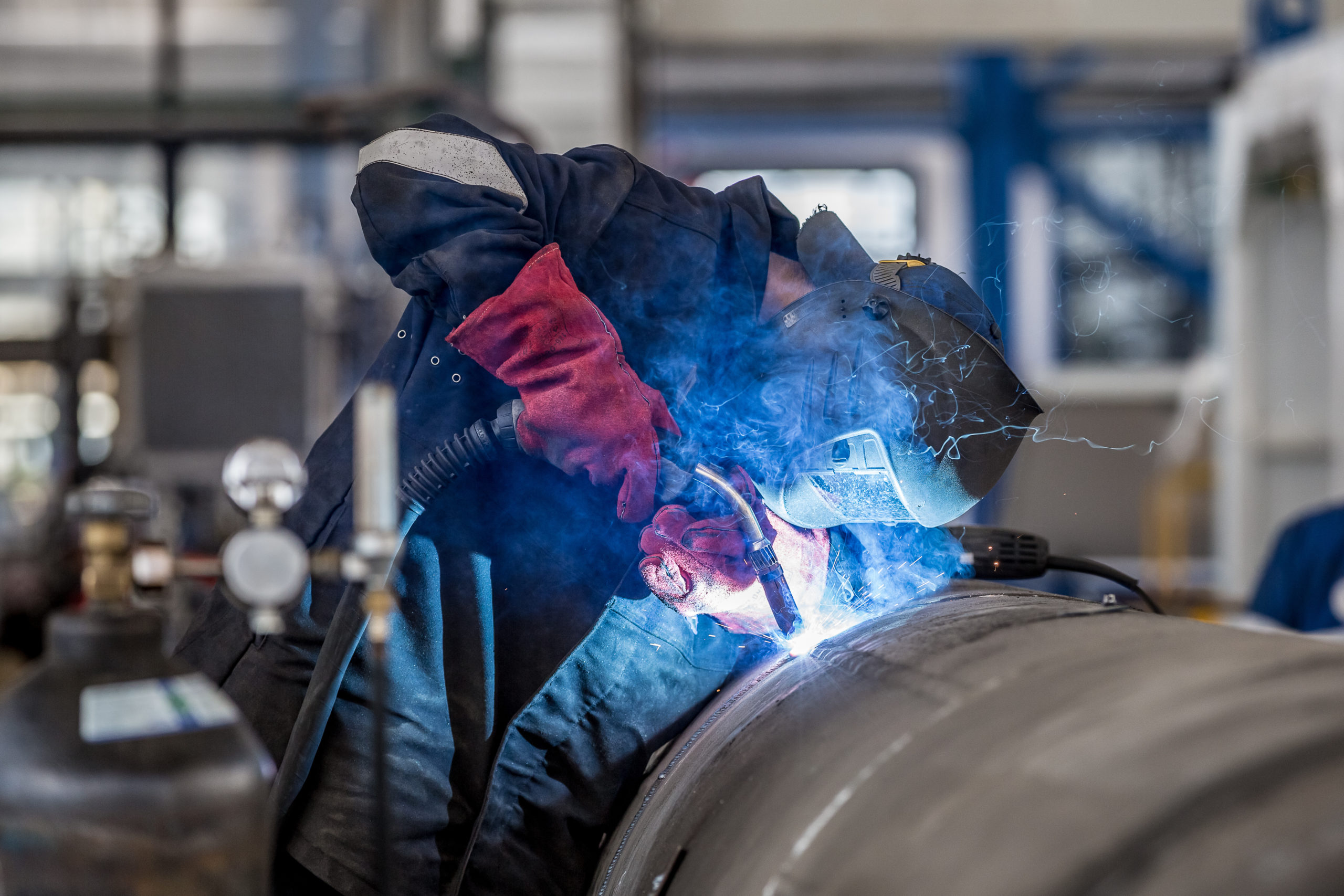 Ein Metallarbeiter in Schutzkleidung und Schweißmaske schweißt eine Naht an einem großen Industrierohr, wobei heller Funkenflug und blaues Licht entstehen.