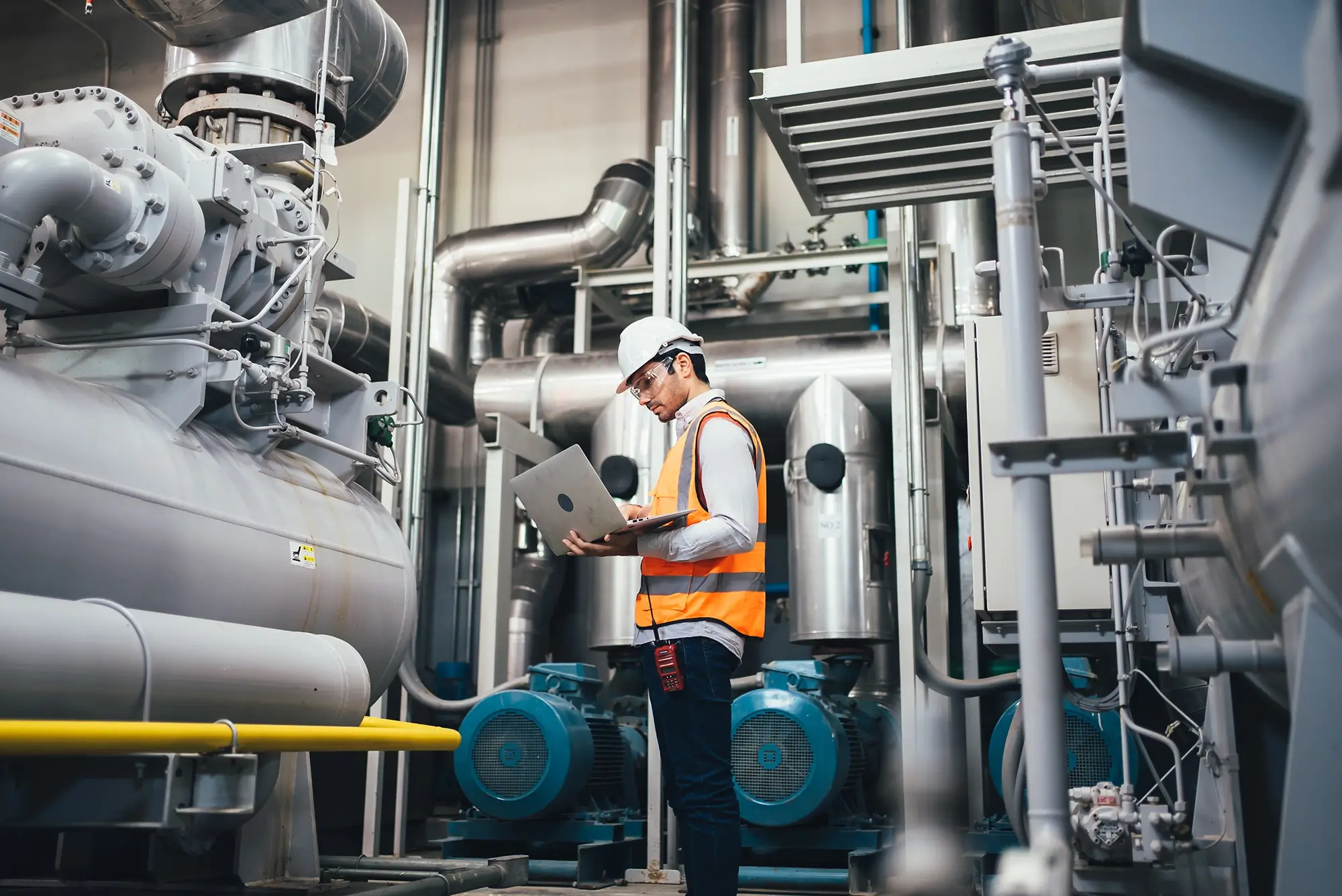 Ein Ingenieur mit weißem Helm und Warnweste arbeitet an einem Laptop inmitten einer Industrieanlage mit großen Maschinen, Pumpen und silbernen Rohrleitungen.