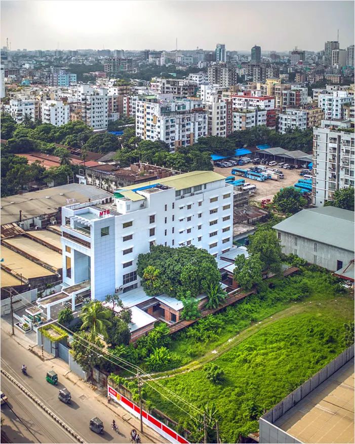 Urban cityscape with mid-rise buildings, a white building with 'MTB' signage, green trees, and parked blue buses in a lot.