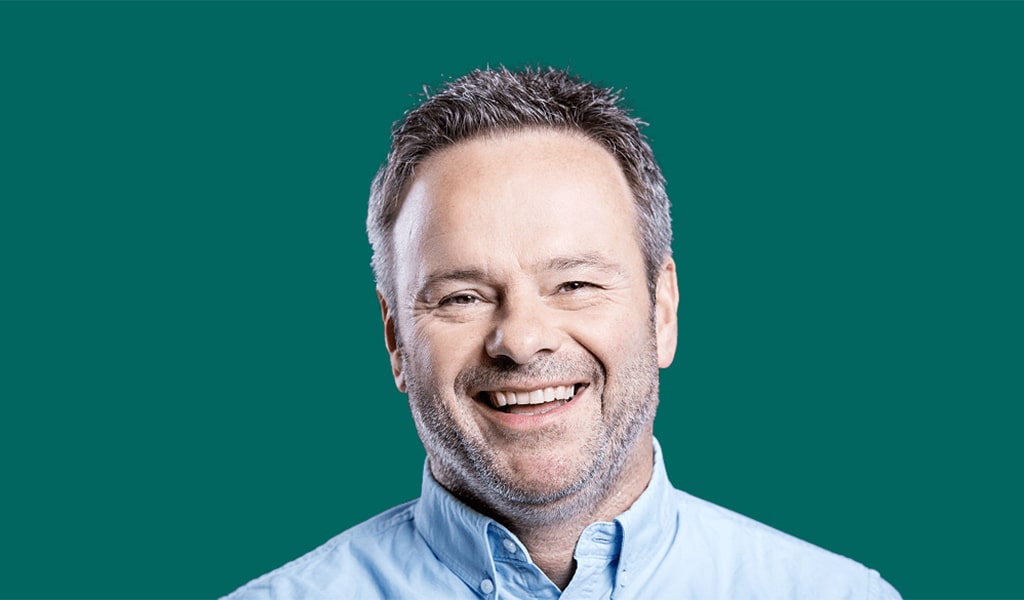 Ein Mann vor einem grünen Hintergrund, der in die Kamera lächelt. 