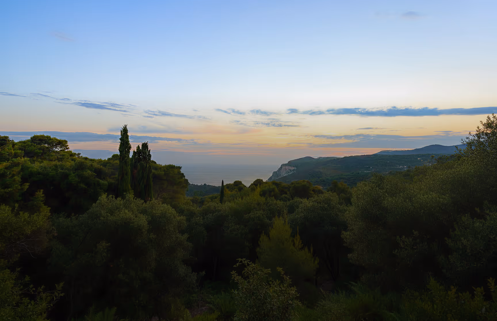 Blick auf bewaldete Hügel mit hohen Zypressen und einem Meer am Horizont bei Sonnenuntergang.