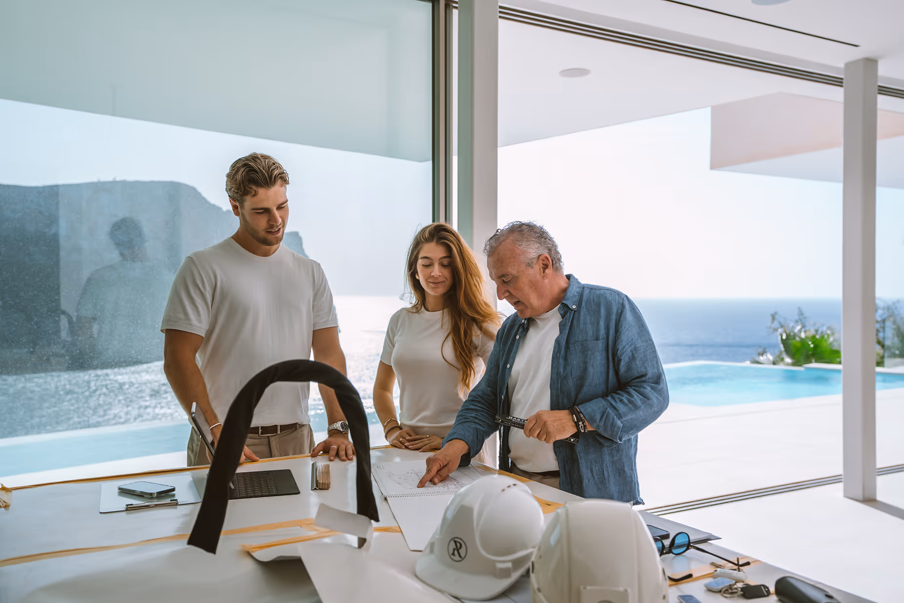 Drei Personen besprechen Baupläne in einem modernen, lichtdurchfluteten Raum mit Meerblick und Pool.