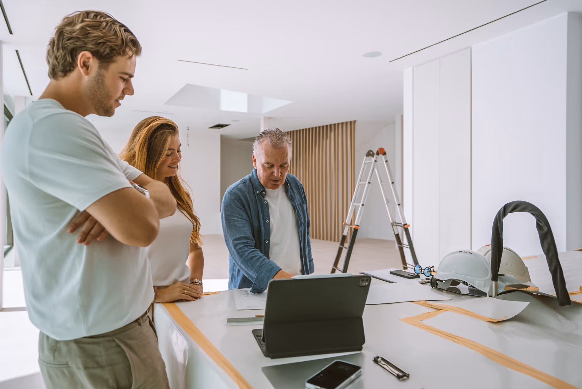 Drei Personen besprechen Baupläne an einem gedeckten Tisch in einem hellen, modernen Raum mit Bauhelmen und einer Leiter im Hintergrund.