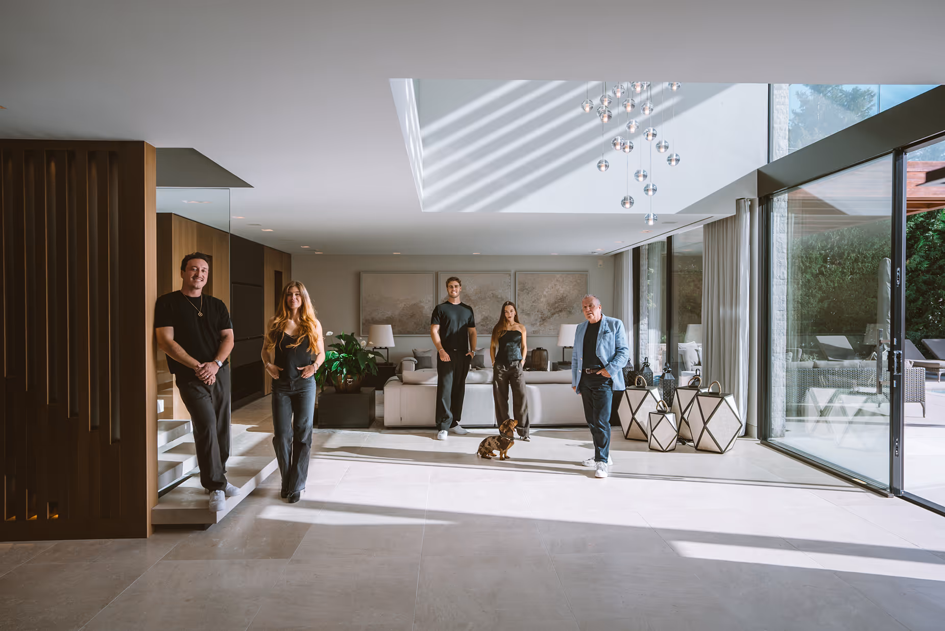 Fünf Personen und ein Hund in einem modernen, hellen Wohnzimmer mit großen Fenstern und einer modernen Lüster.