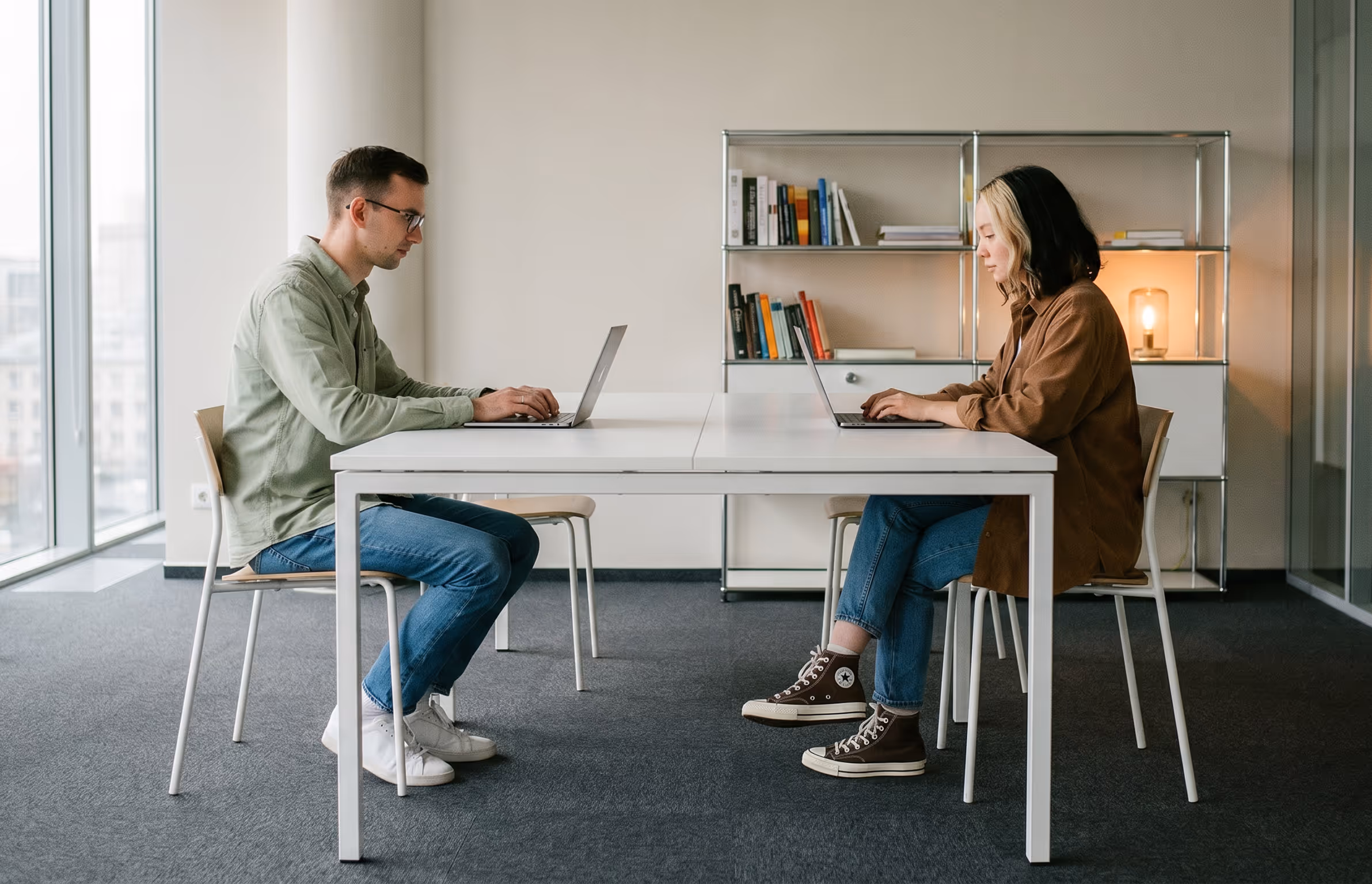 Man and woman sitting opposite each other at a white table working on laptops in a modern office with a bookshelf and lamp in the background.