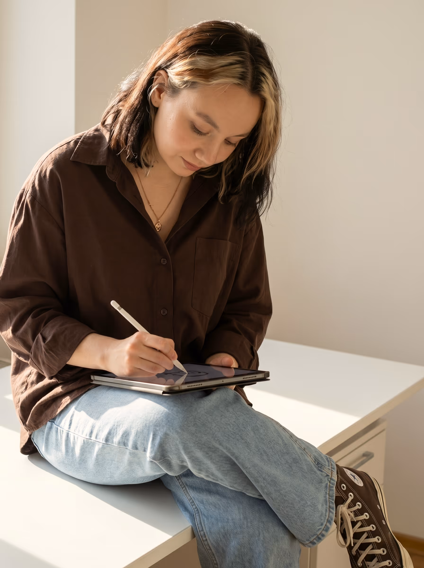 Young woman with dark and blonde hair wearing a brown shirt and jeans, drawing on a tablet with a stylus while sitting on a white desk.