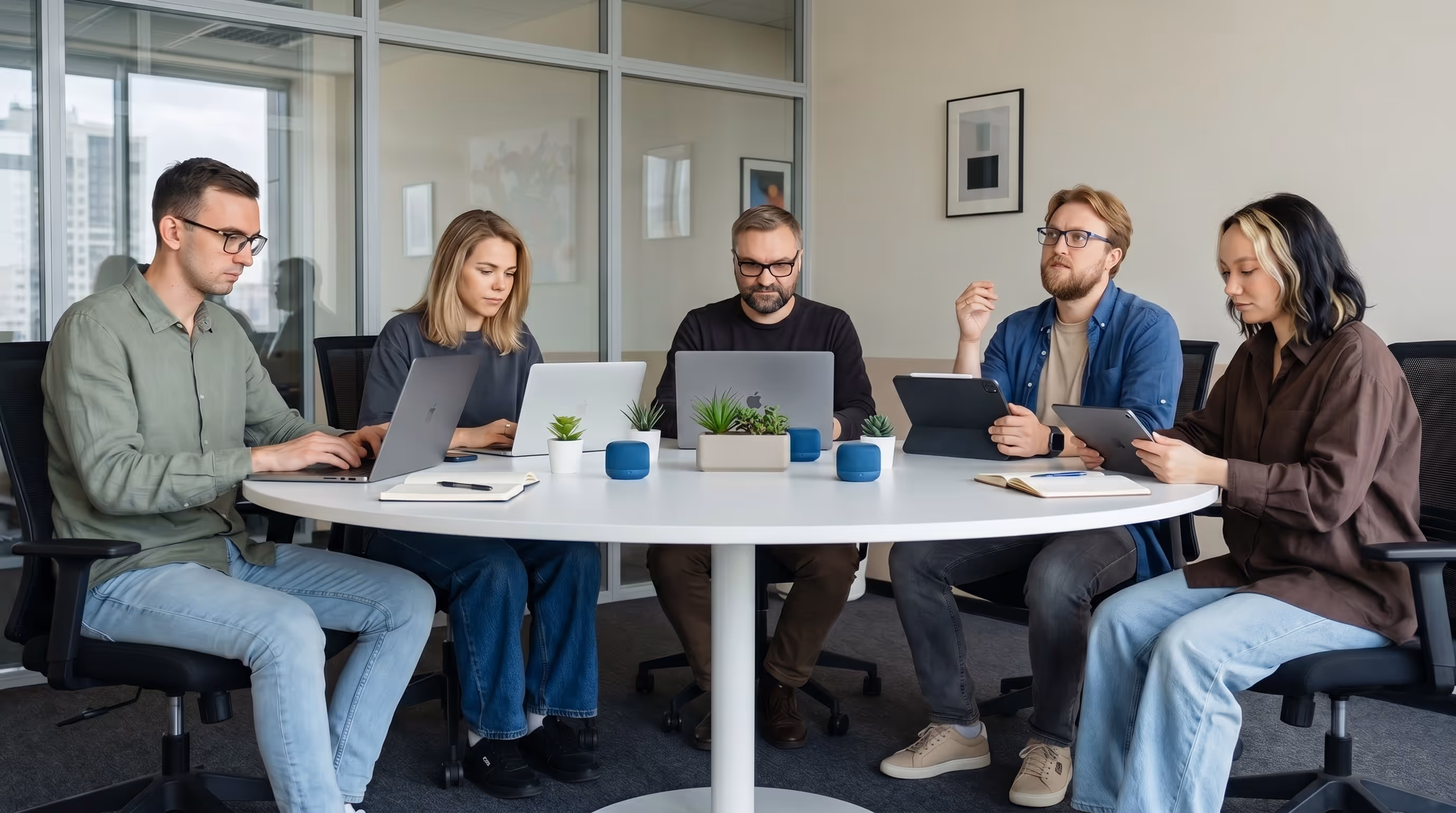 Five young professionals working on laptops and tablets around a round white table with small plants and blue speakers in a modern office.
