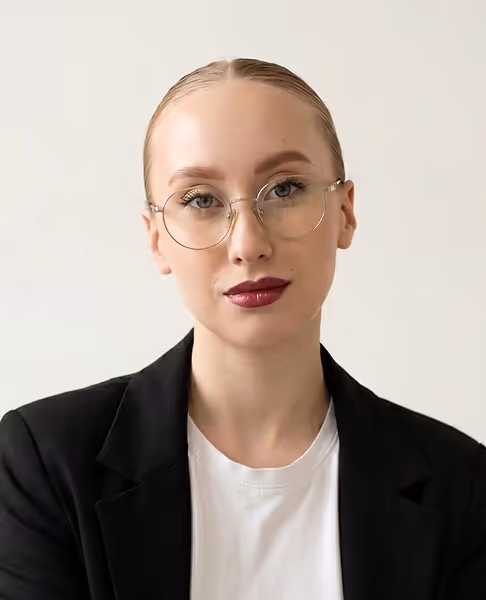 Young woman with slicked-back hair, wearing round glasses, dark lipstick, a black blazer, and white shirt against a plain light background.
