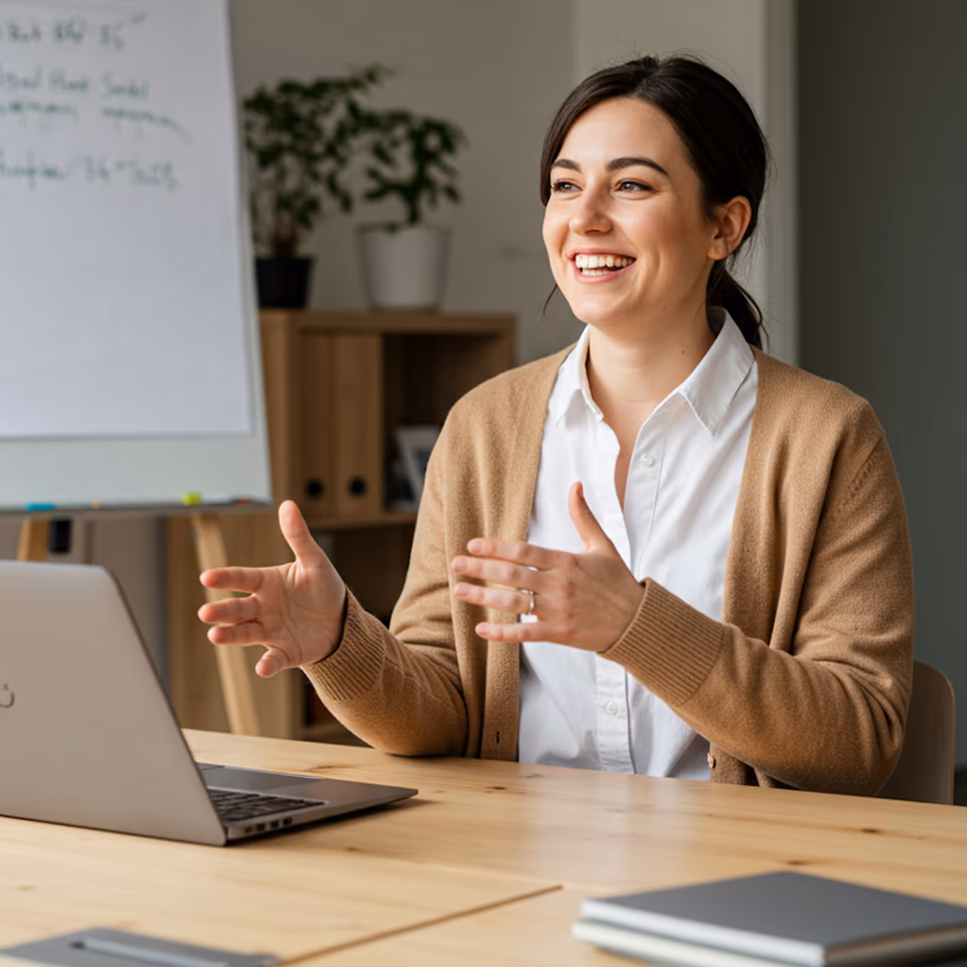 Lächelnde Frau in brauner Strickjacke spricht vor einem Laptop in einem Büro mit Whiteboard und Pflanzen im Hintergrund.