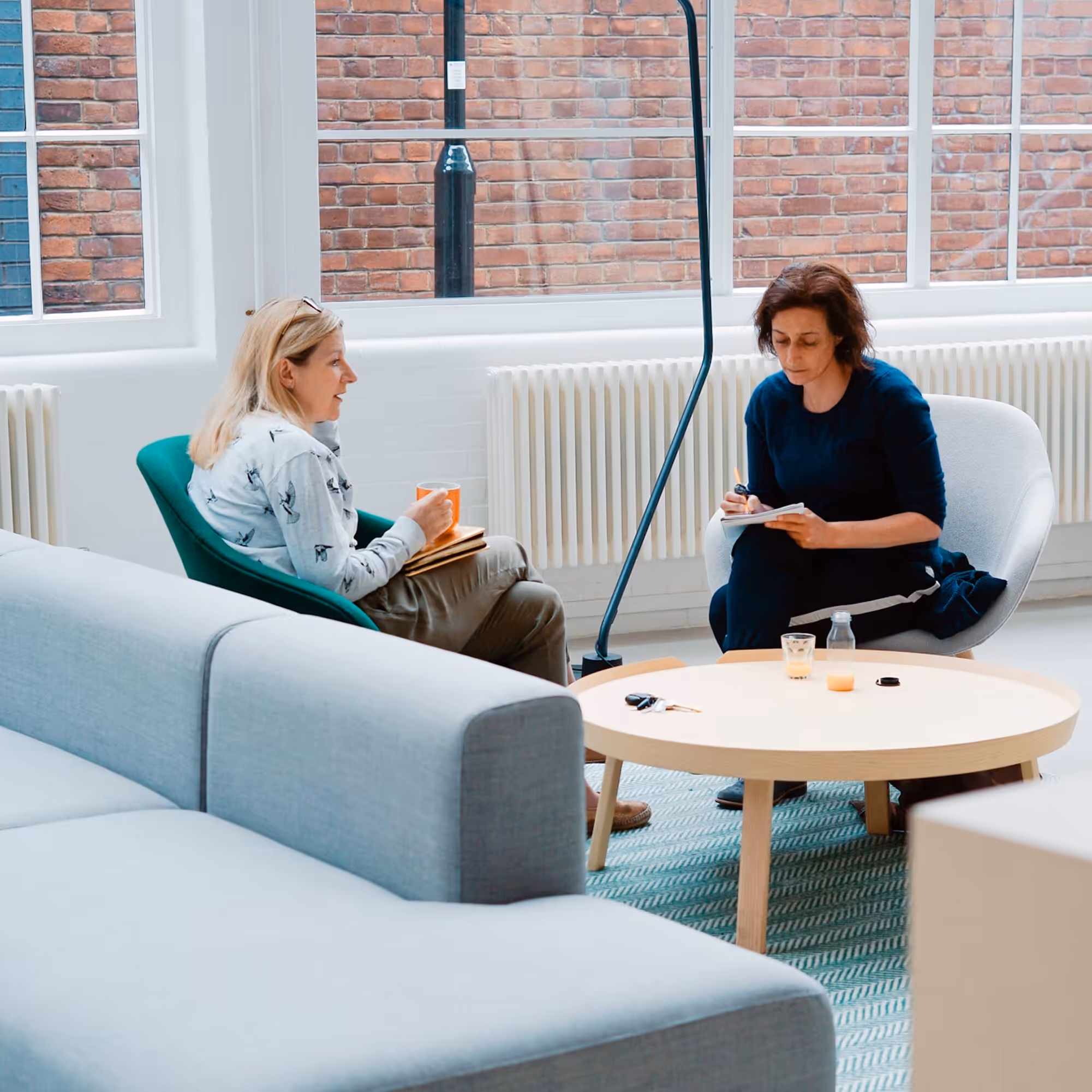 Zwei Frauen sitzen in einem hellen Raum mit großen Fenstern gegenüber und führen ein Gespräch, eine hält eine Tasse, die andere macht Notizen.