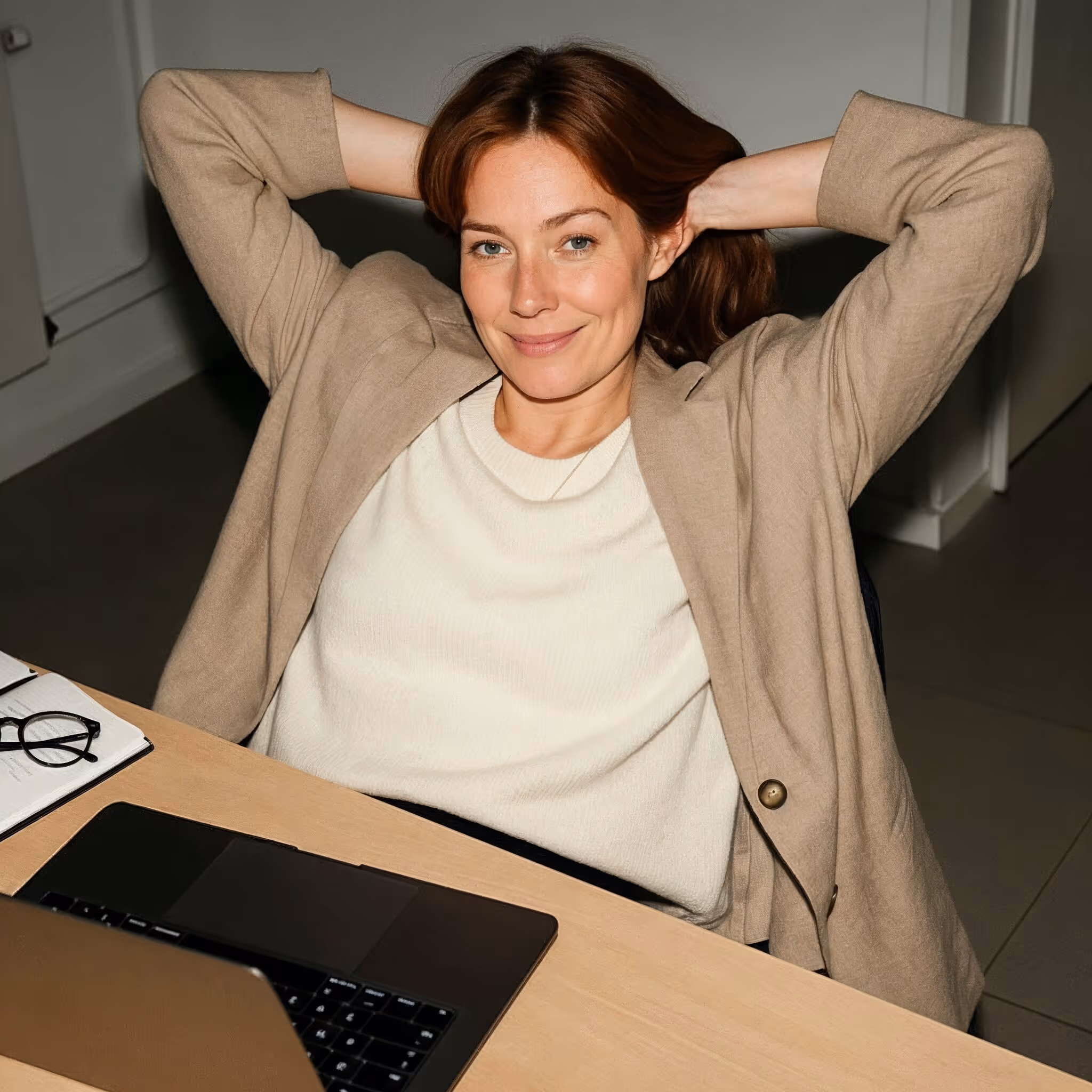 Lächelnde Frau in einem beigen Blazer, die sich mit den Händen hinter dem Kopf an einem Schreibtisch mit Laptop und Brille zurücklehnt.