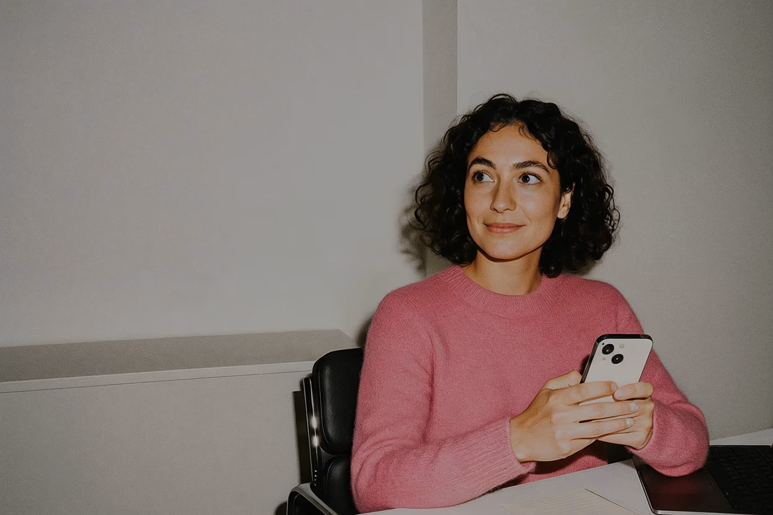 Frau mit lockigem Haar und pinkem Pullover hält ein Smartphone und schaut lächelnd zur Seite.