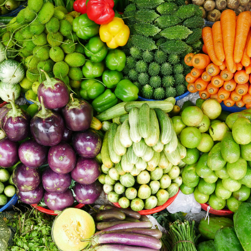 Une variété de légumes frais colorés entassés, dont des aubergines violettes, des poivrons verts, rouges et jaunes, des concombres, des carottes orange, des citrons verts, des melons coupés et d'autres légumes verts.