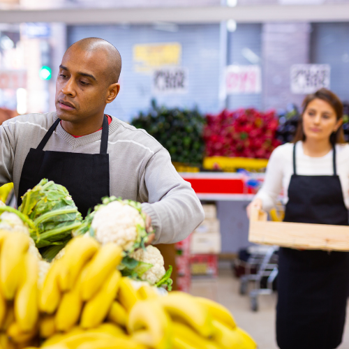 Un homme portant un tablier noir organise des légumes, dont du chou-fleur, dans un marché ou une épicerie, tandis qu'une femme en arrière-plan tient une caisse.