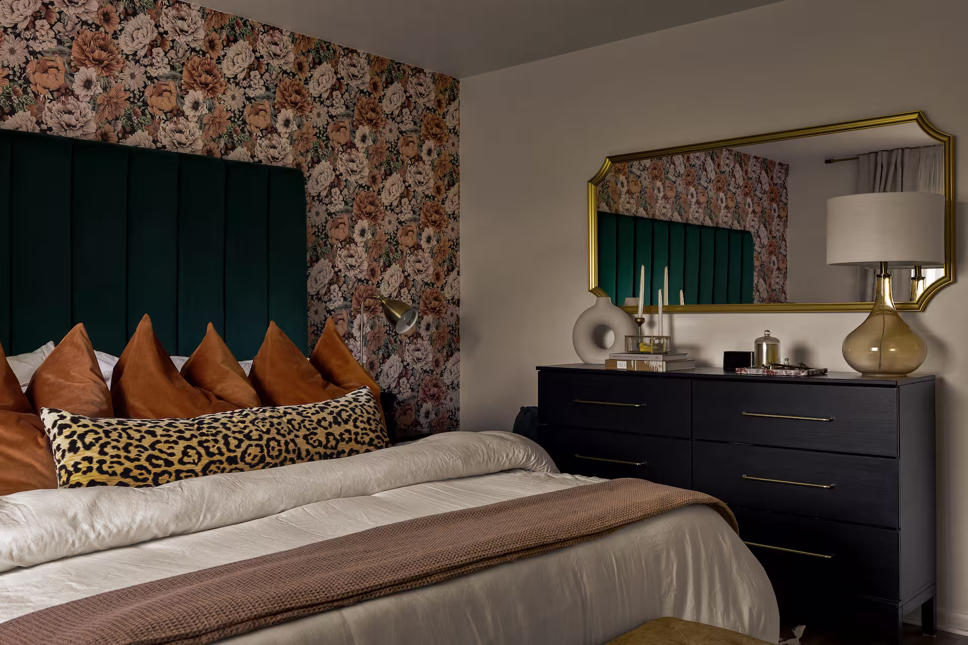 Cozy bedroom with floral wallpaper, a dark green upholstered headboard, brown and leopard print pillows, and a black dresser topped with a gold-framed mirror and decorative items. | Jade Mystic Interiors