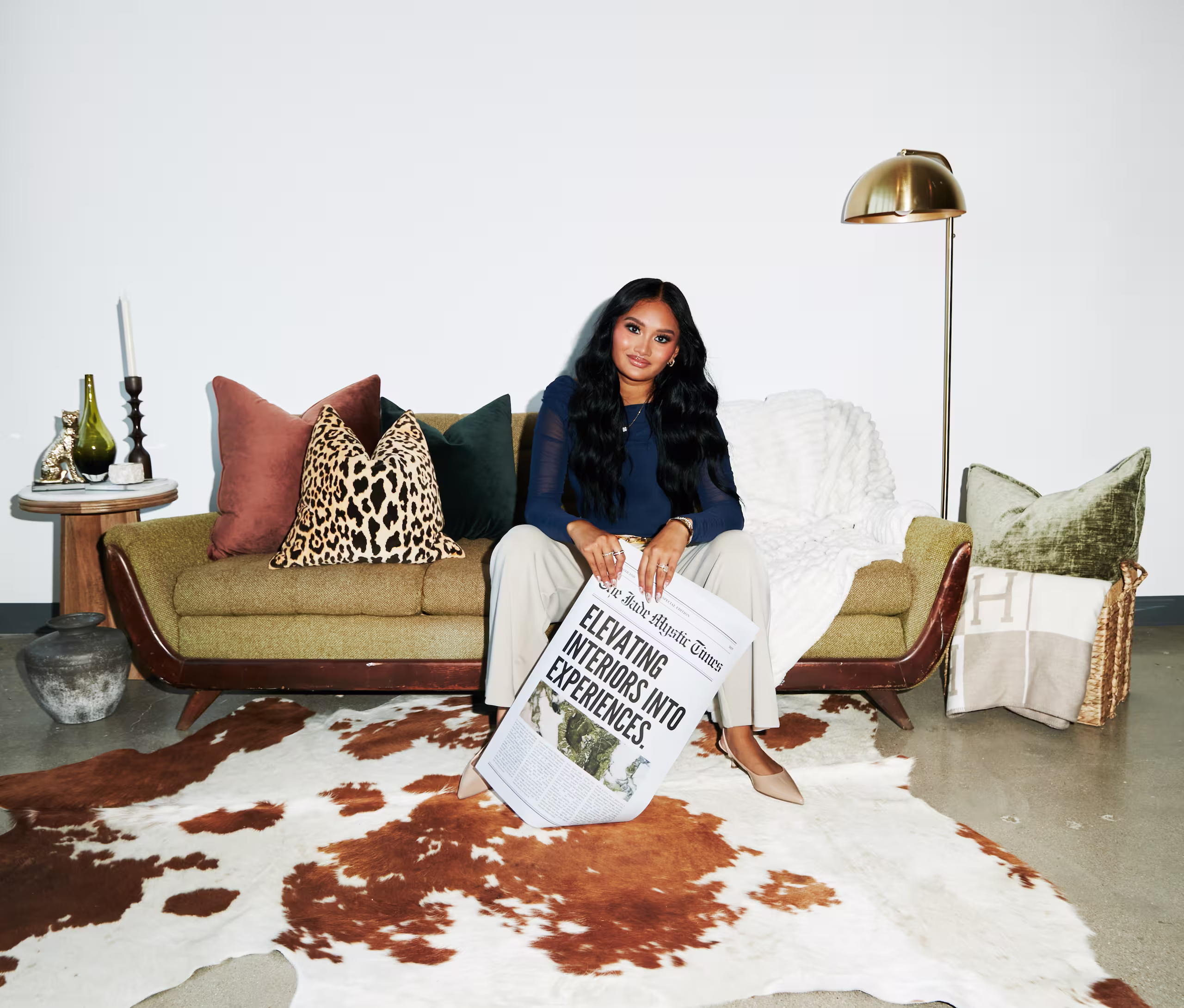 Woman sitting on a green couch with patterned pillows holding a newspaper titled 'Elevating Interiors Into Experiences' on a brown and white cowhide rug.