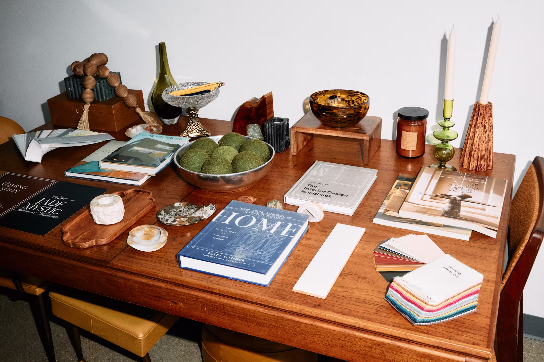 Wooden table with interior design books, green decorative balls in a metal bowl, candles, color swatches, and various decorative objects.