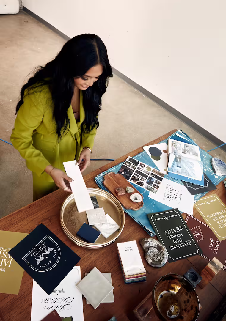 Woman in a green jacket examining fabric swatches and design materials on a wooden table. | Jade Mystic Interiors