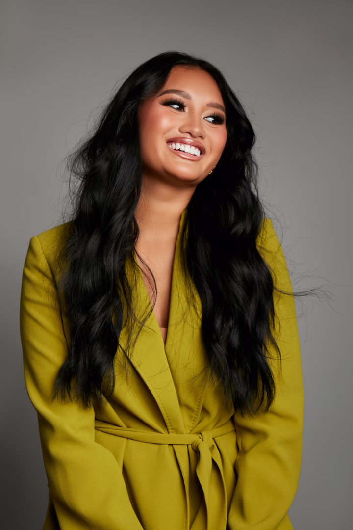 Smiling woman with long black wavy hair wearing a mustard-yellow belted blazer against a plain gray background. Jade Mystic Interior Aesthetics