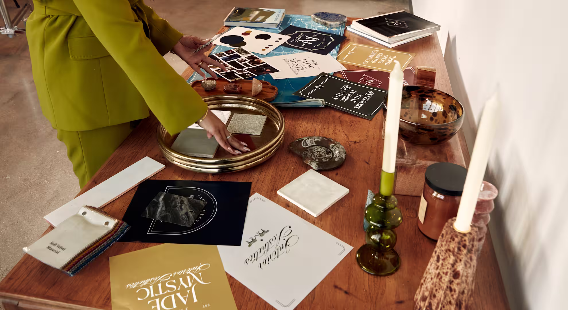 Person in a green suit arranging decorative items and design materials on a wooden table, including candles, books, and sample cards. Jade Mystic Interior Aesthetics