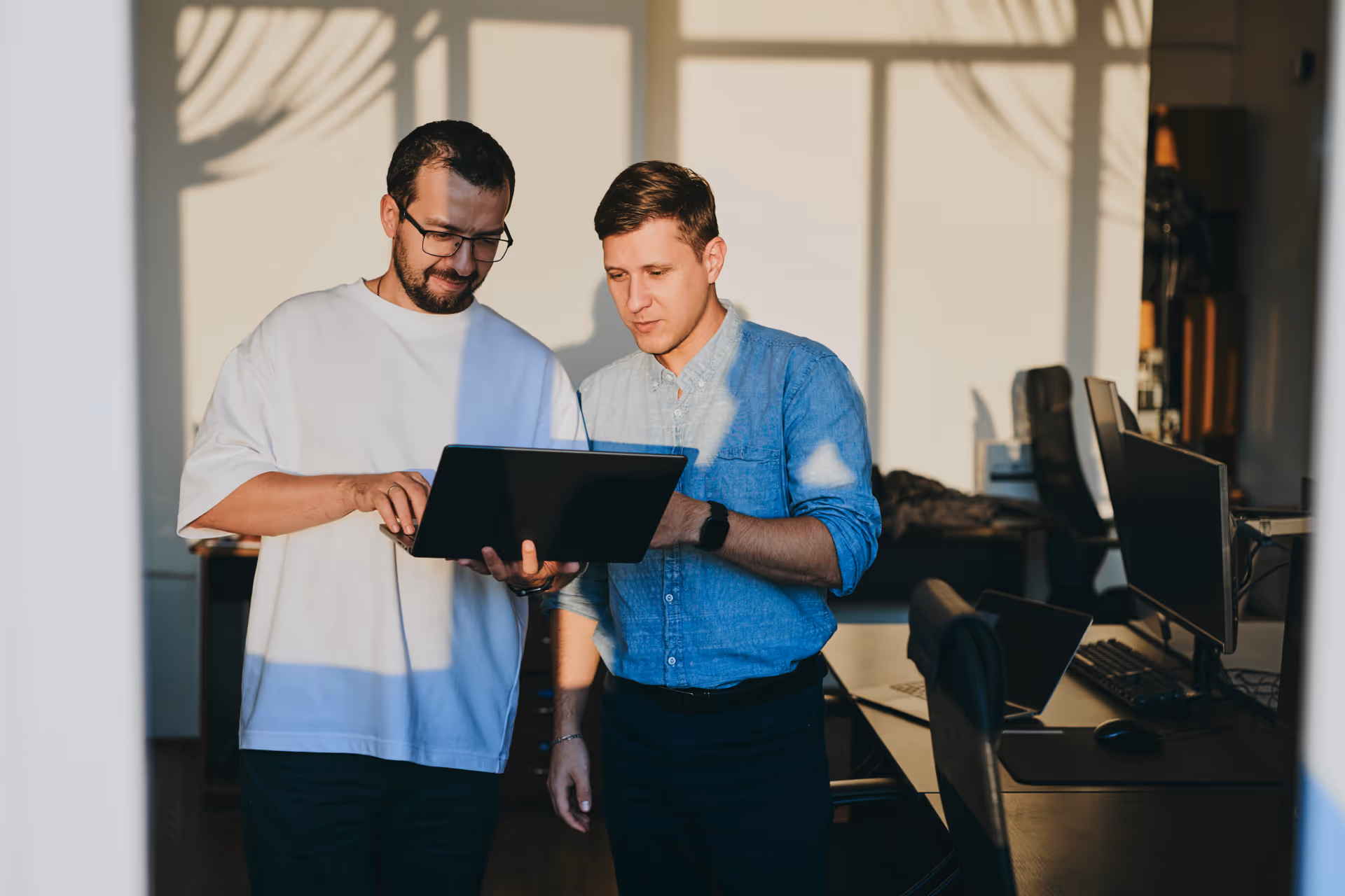 Two men standing side by side in an office, looking at a laptop screen together.