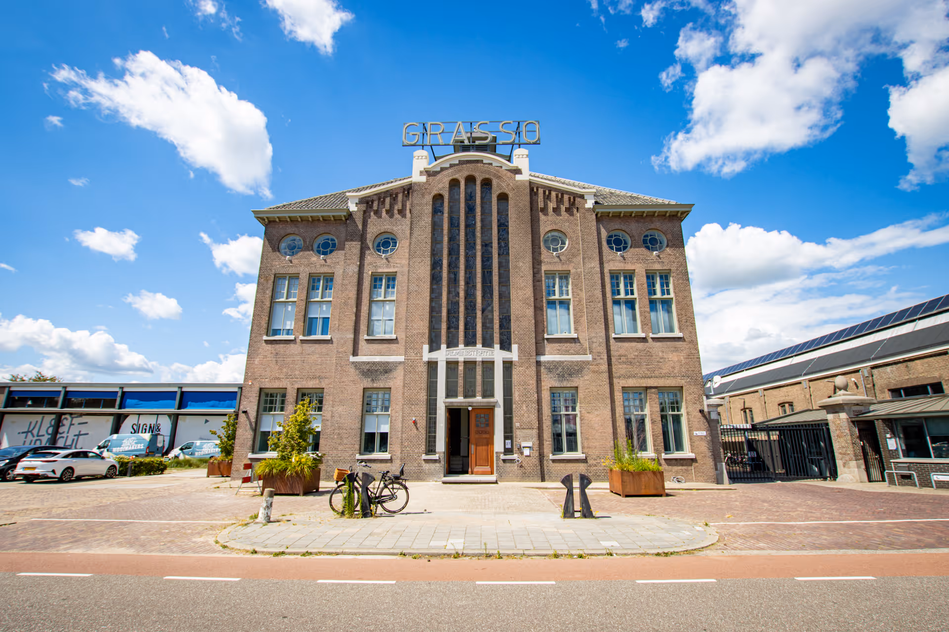 Historic brick building with tall vertical windows and a sign reading 'GRASSI' on the roof, under a partly cloudy blue sky.