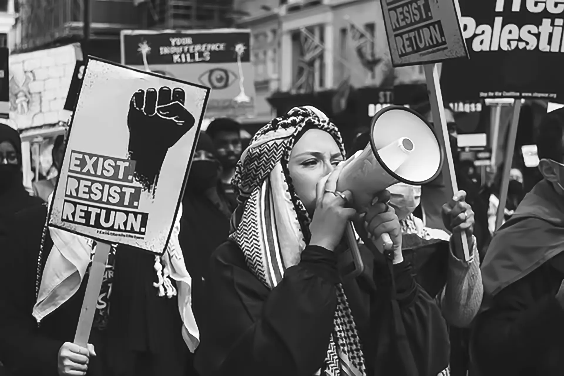 Person wearing a keffiyeh speaking into a megaphone during a protest with signs reading 'Exist. Resist. Return.' and 'Free Palestine.'