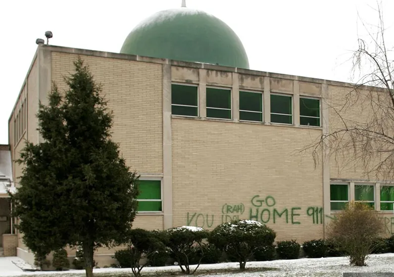 Exterior of a beige brick mosque with a green dome, showing graffiti with hate speech on the wall.