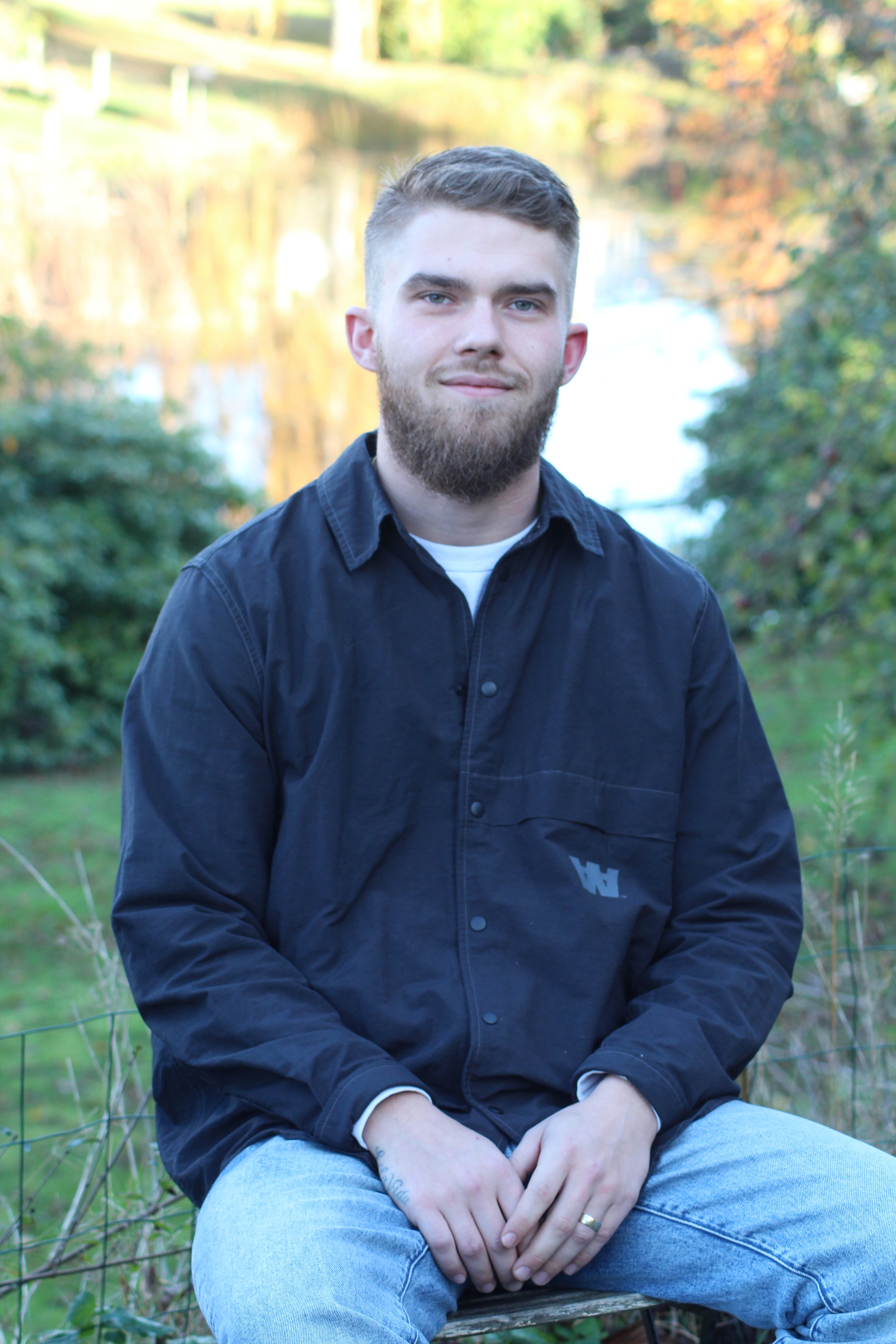 Young man with beard sitting outdoors on a bench wearing a black jacket and light blue jeans.