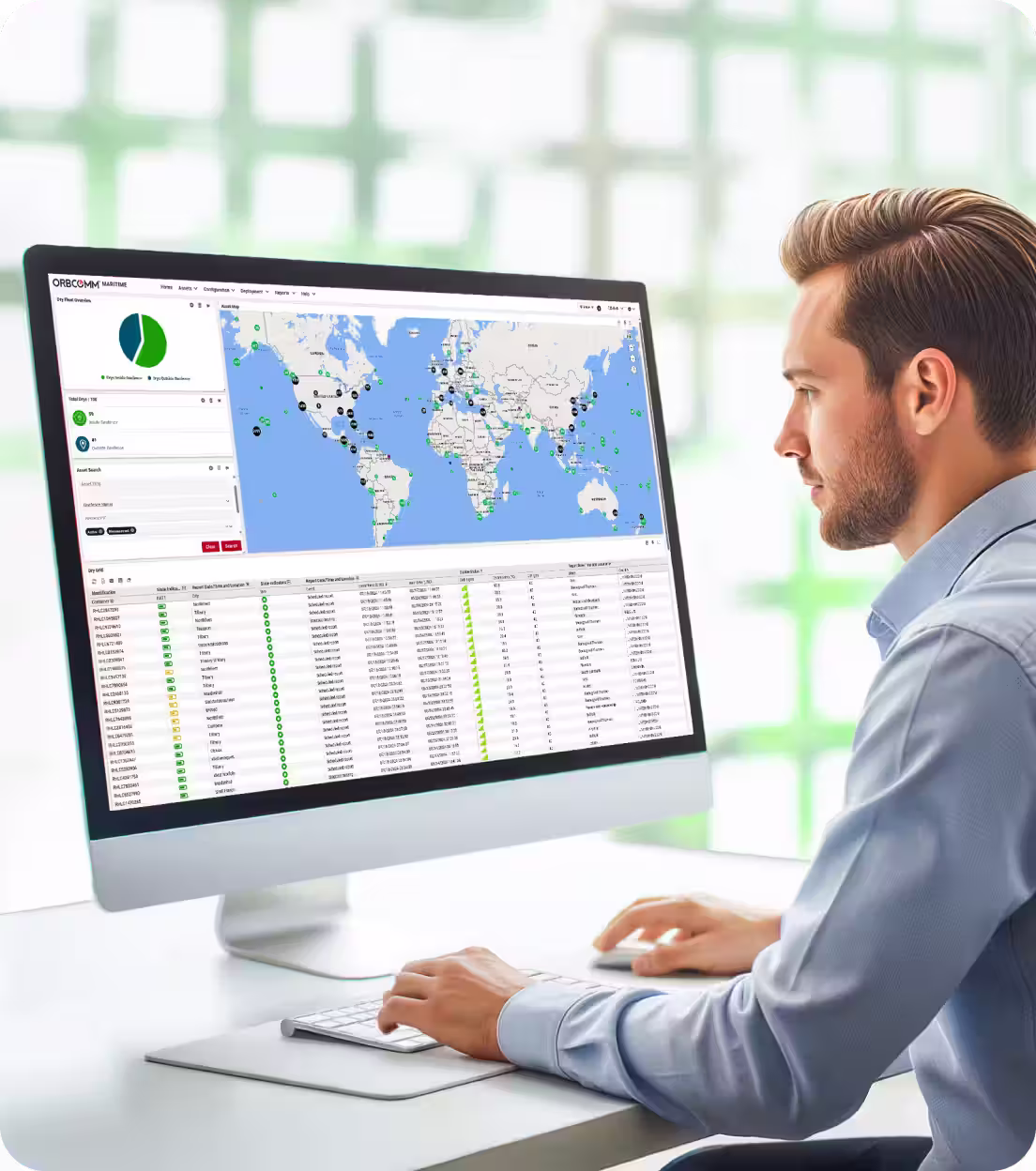 Man in blue shirt using a computer displaying a global map and data dashboard for asset tracking and dry fleet management.