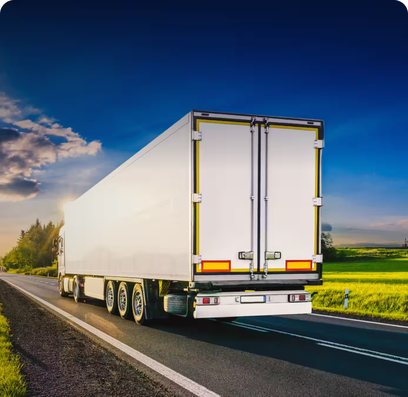 White cargo truck driving on a highway beside green fields under a clear blue sky at sunset.