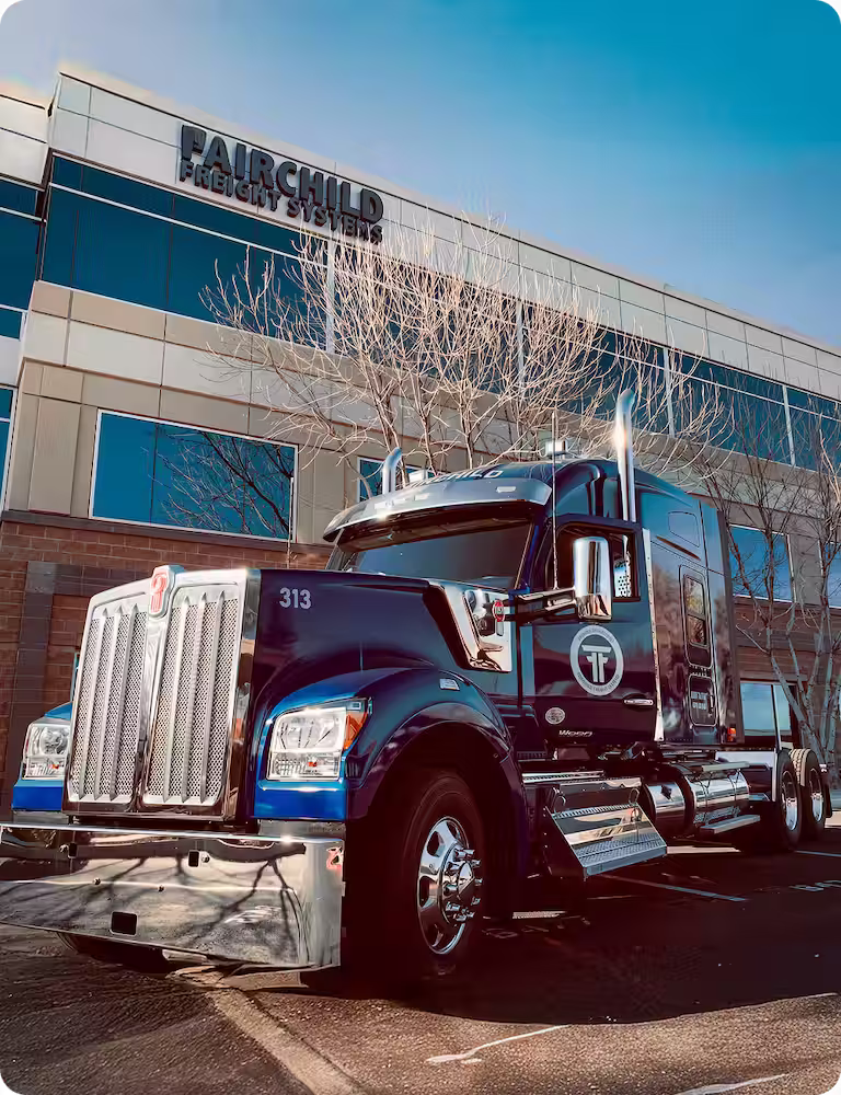 Large blue and chrome semi-truck parked in front of a building with Fairchild Freight Systems signage, under a clear blue sky.