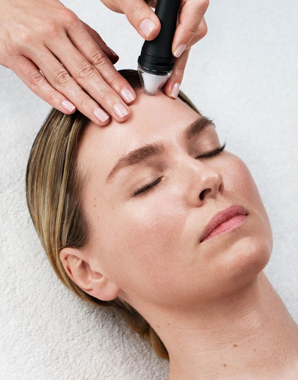 Kosmetikerin führt eine Gesichtsbehandlung mit einem Handgerät auf der Stirn einer entspannten Frau durch.