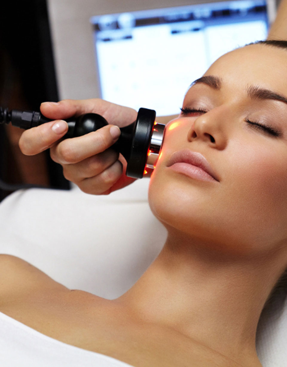 Kosmetikerin führt eine Radiofrequenz- oder LED-Gesichtsbehandlung durch; ein Gerät mit rot leuchtenden Lichtquellen wird sanft auf die Wange einer entspannten Frau gehalten.