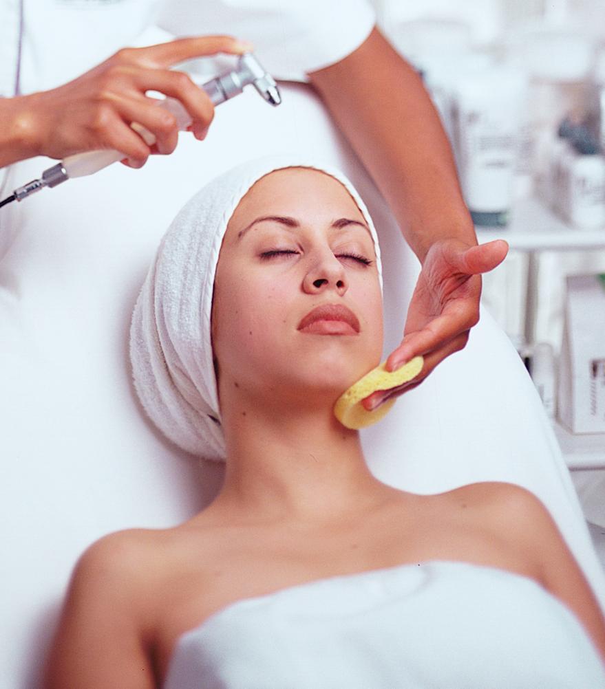 Kosmetikerin führt eine Gesichtsbehandlung durch; eine Frau liegt entspannt mit Handtuchturban auf der Liege, während ihr Hals mit einem Schwamm gereinigt wird.