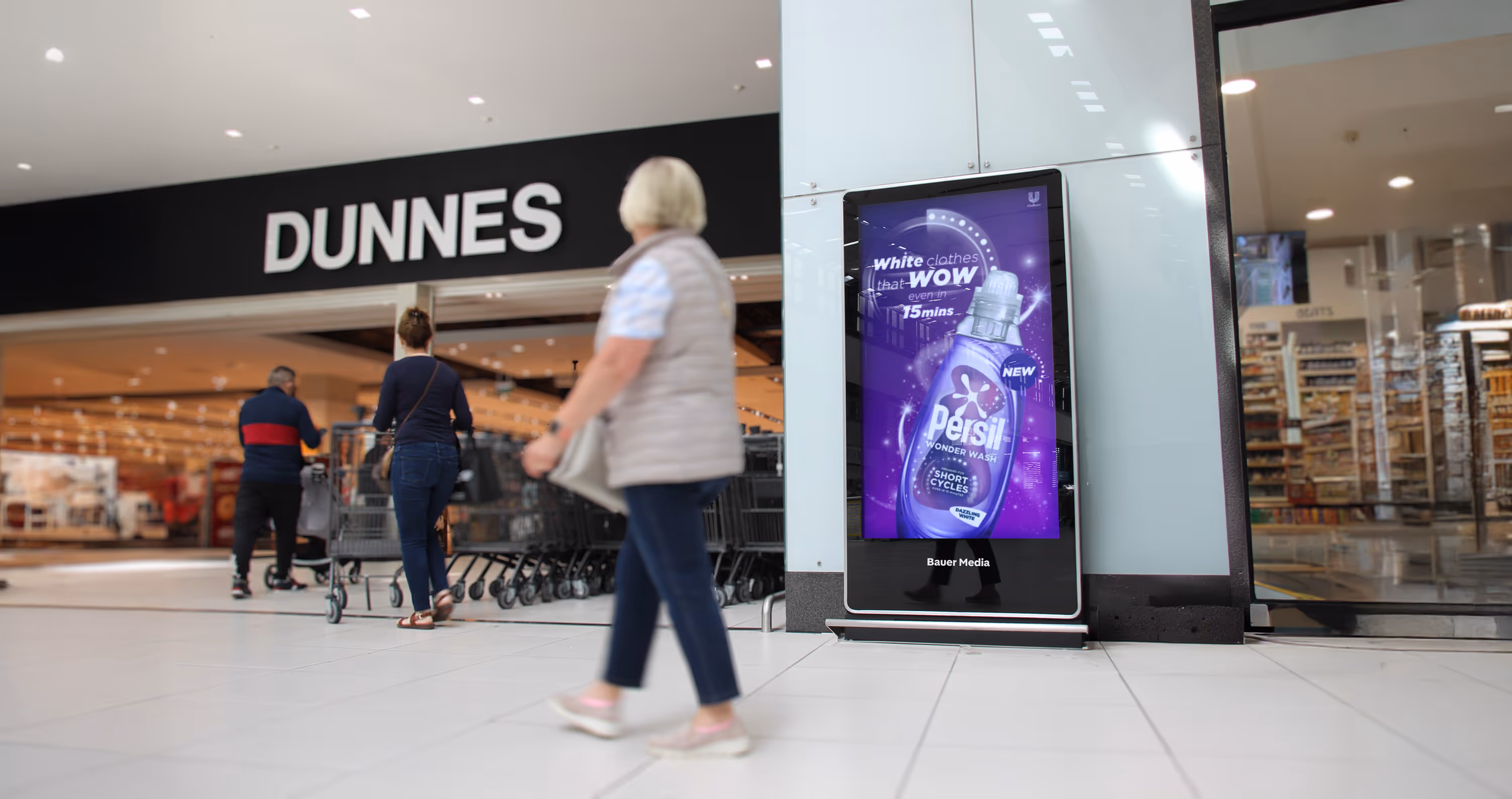 An Adshel Live Retail digital screen displaying an advertisement for Heinz Mayonnaise, situated outside the entrance of a Tesco supermarket in a shopping mall environment