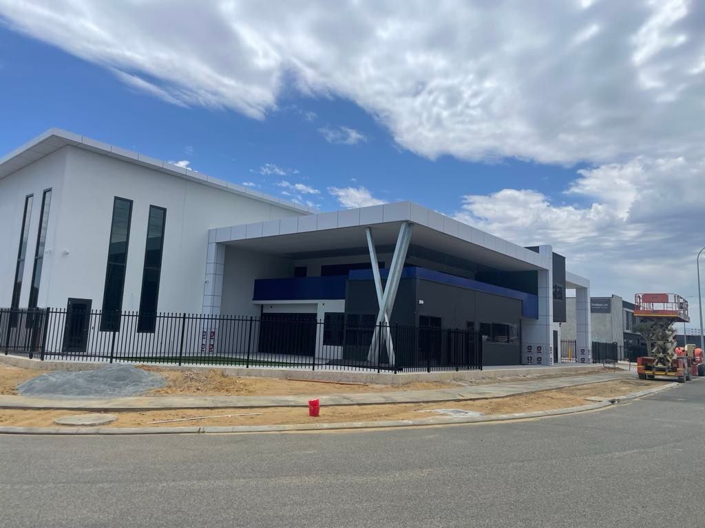 Modern industrial building with white and black exterior, blue accents, and a fenced perimeter under a partly cloudy sky.