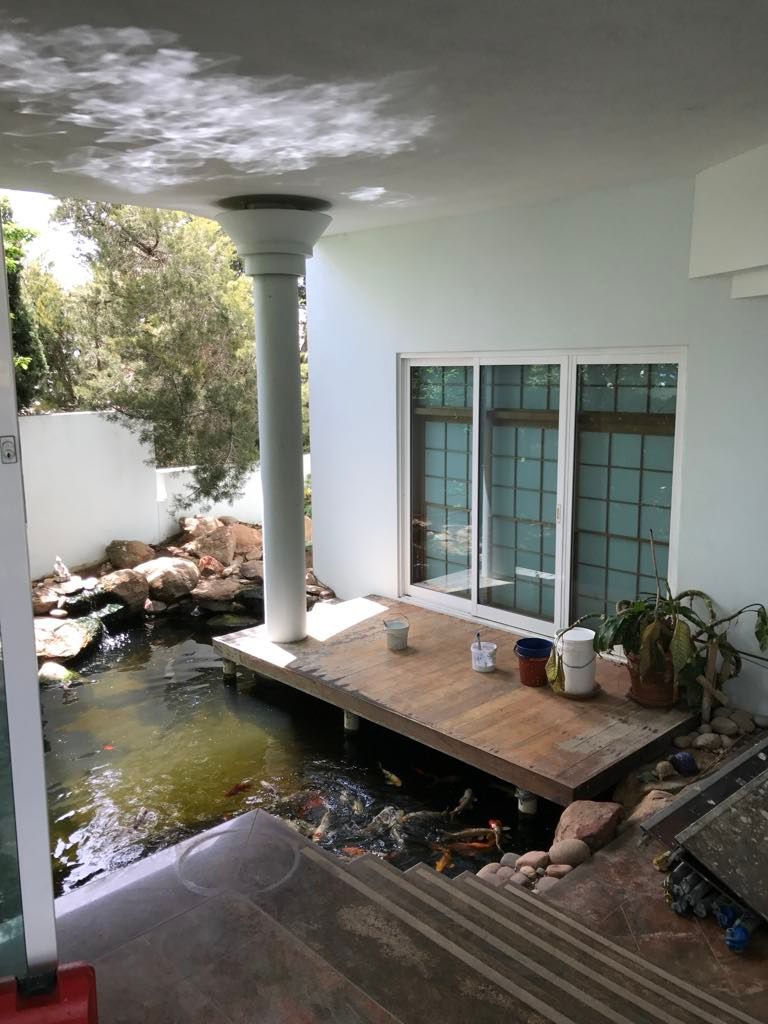 Outdoor koi pond with large rocks, a wooden deck with buckets and a potted plant, next to sliding glass doors and shaded by a white ceiling with a pillar.