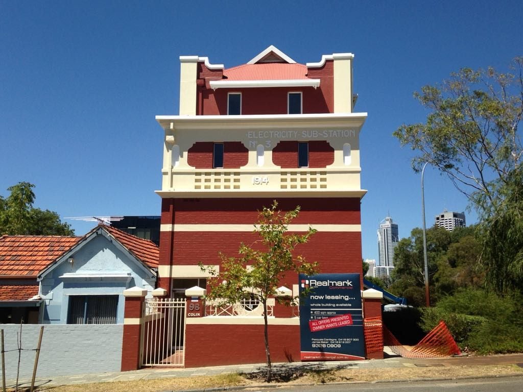 Red and cream historic electricity sub-station building from 1914 with a real estate sign advertising leasing in front under a clear blue sky.