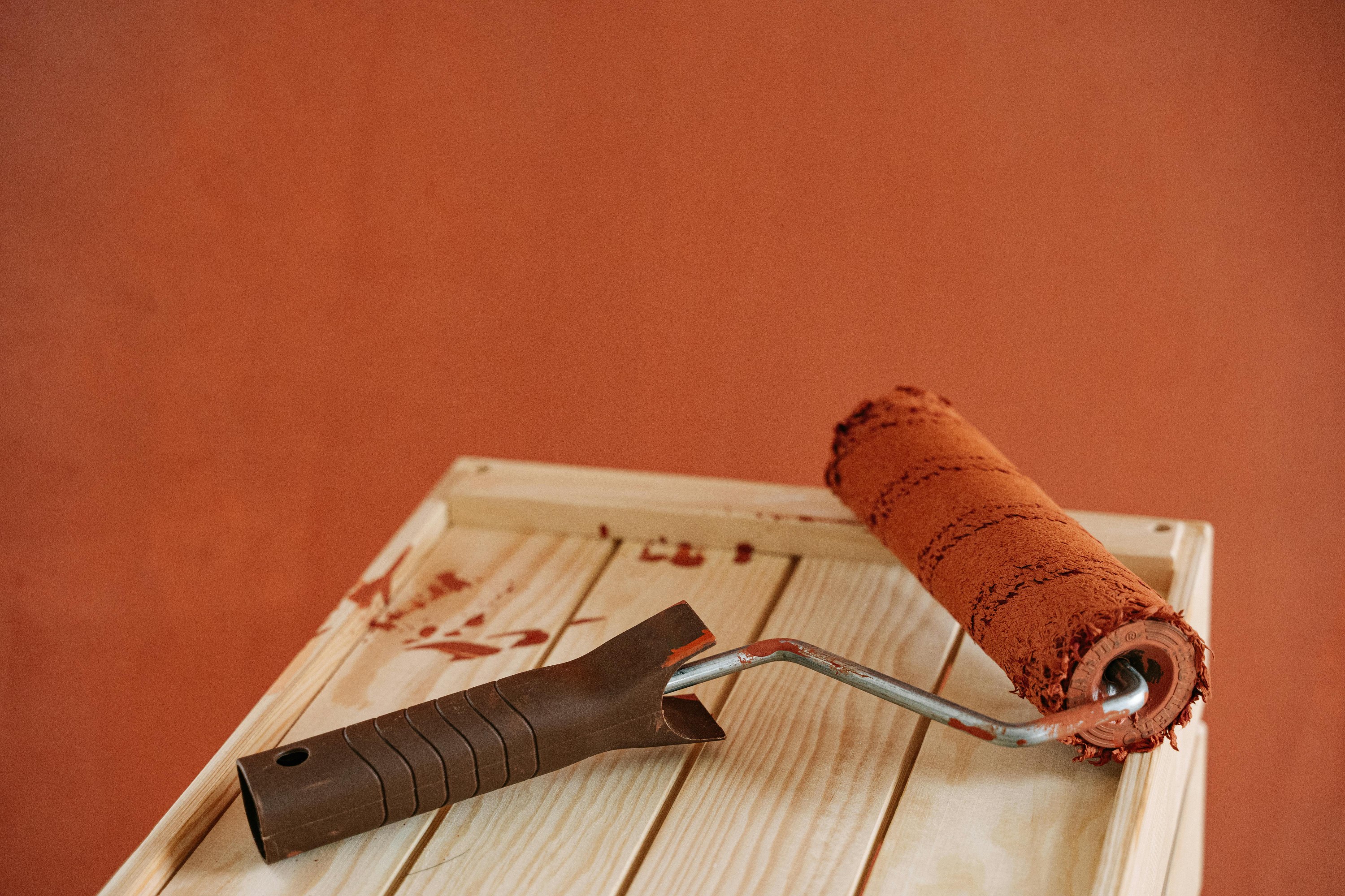 Paint roller with red paint resting on a wooden surface against a red wall.