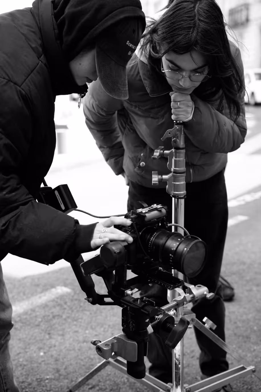 Gros plan en noir et blanc de deux personnes, vêtues de doudounes, se penchant pour régler une caméra professionnelle montée sur un trépied à l'extérieur. L'une d'elles touche les commandes de la caméra.