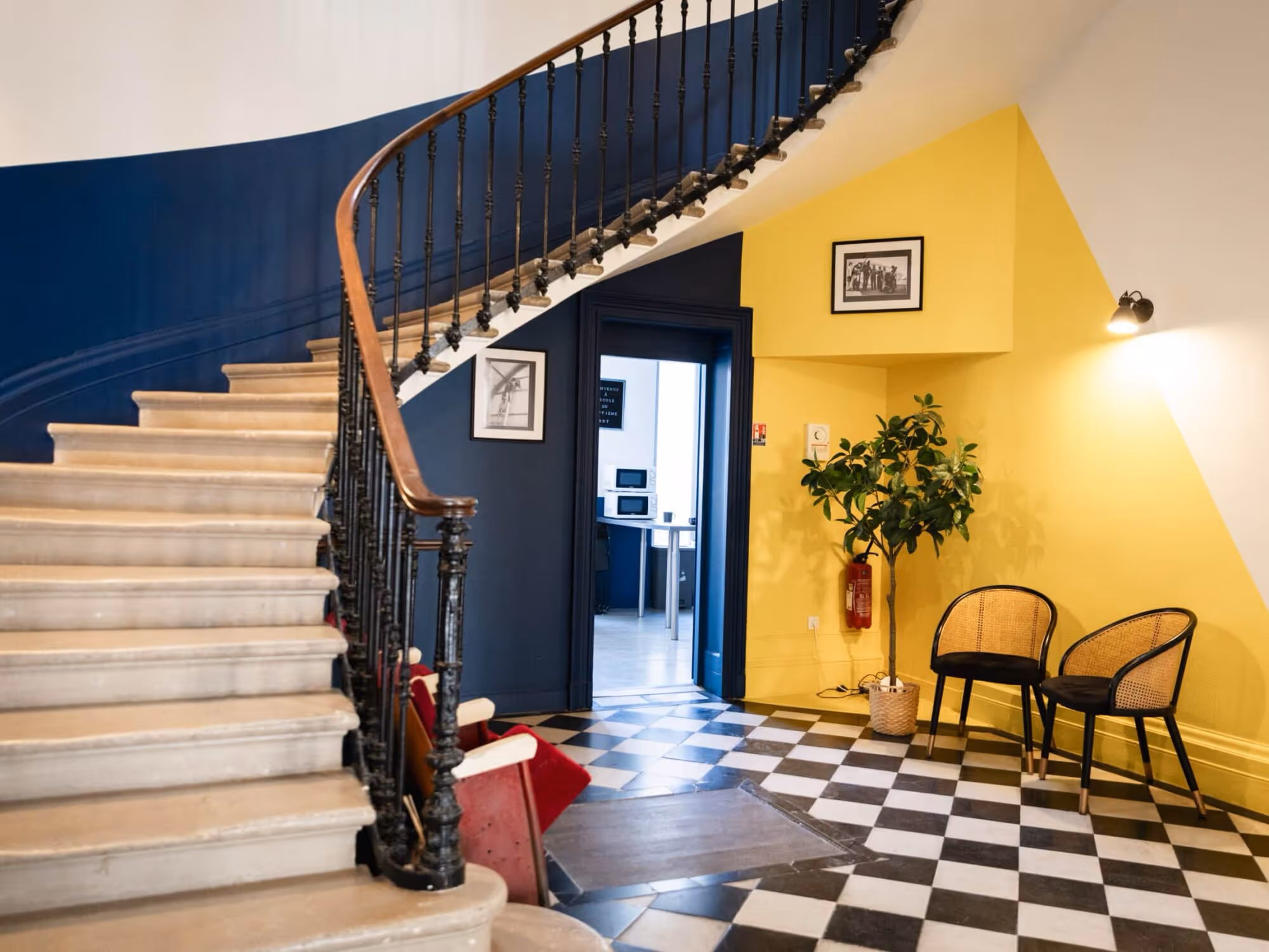 Vue intérieure de l'entrée de l'École du Septième Art, montrant un escalier en pierre avec une rampe en fer forgé, un sol en damier noir et blanc, et des murs peints en jaune vif et bleu marine.