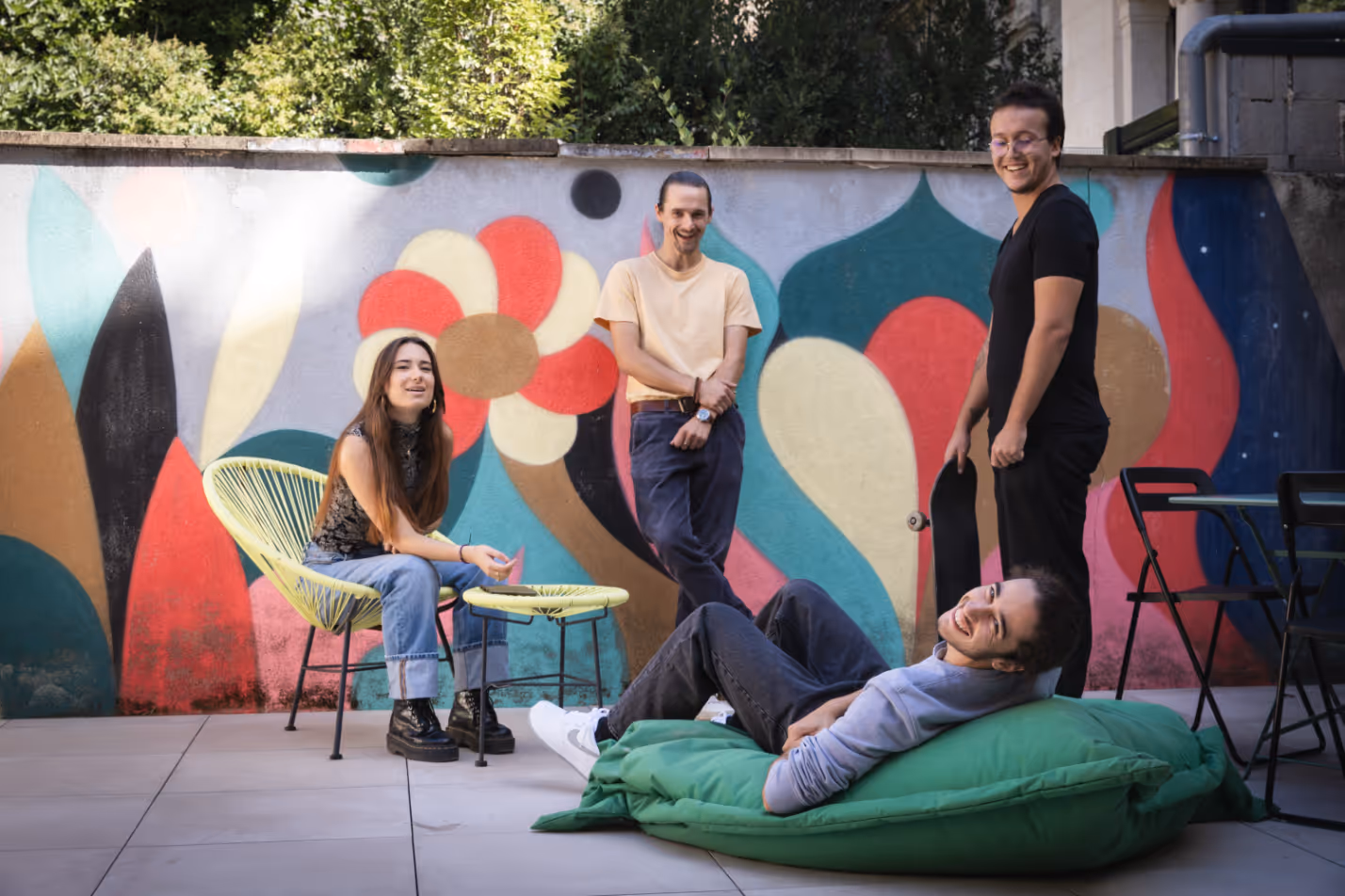 Quatre jeunes étudiants souriants se détendent à l'extérieur, assis sur des poufs et des chaises sur fond de mur avec une fresque colorée et abstraite. L'image représente la vie étudiante décontractée.