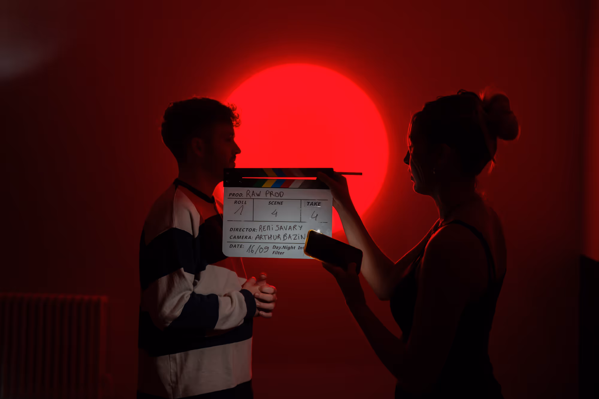 Silhouettes of a man and a woman with a clapperboard in front of a bright red circular light background during a film shoot.