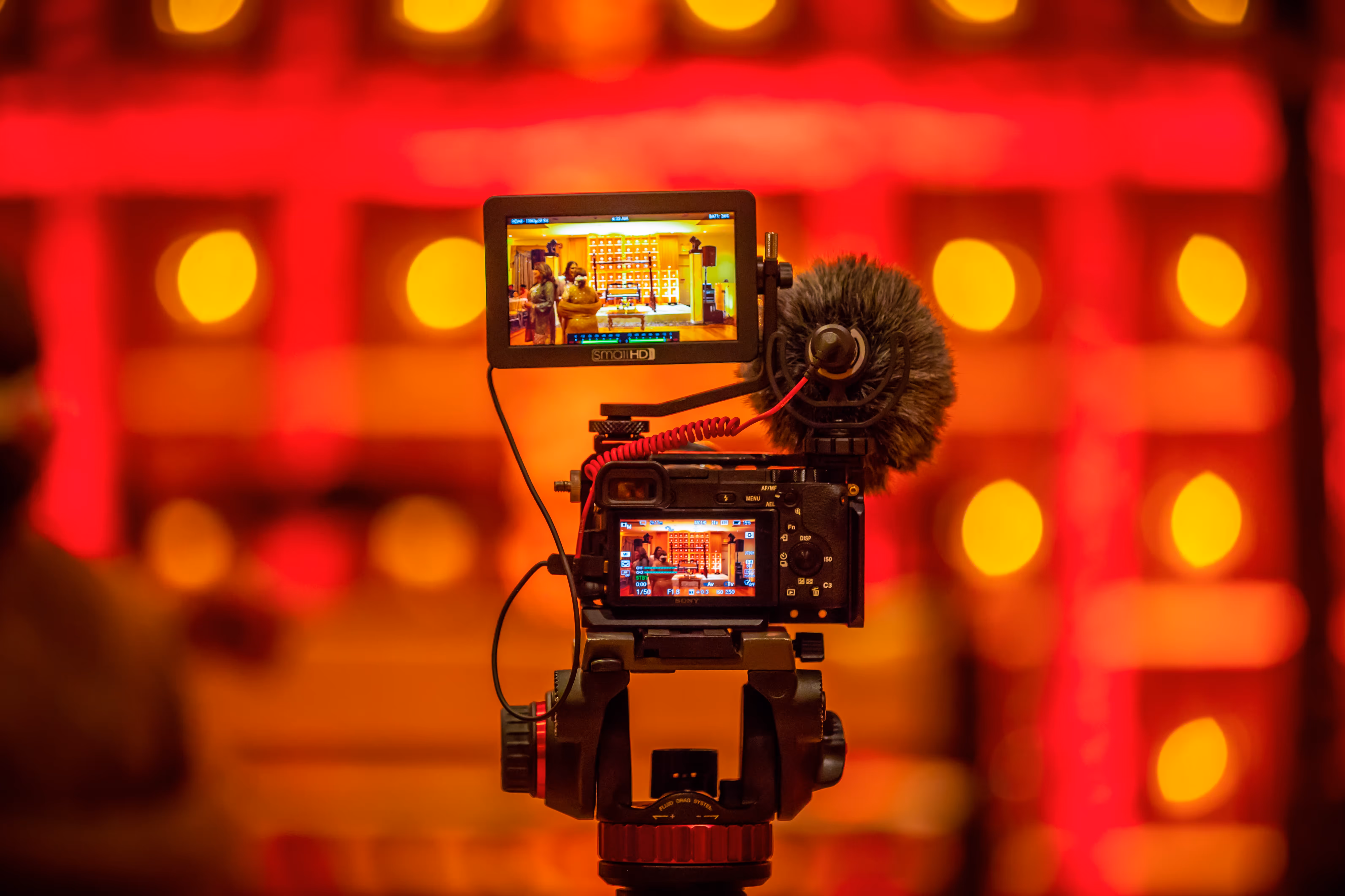 Caméra professionnelle avec micro externe filmant une scène avec deux personnes dans un décor chaleureux aux lumières orange et rouges.