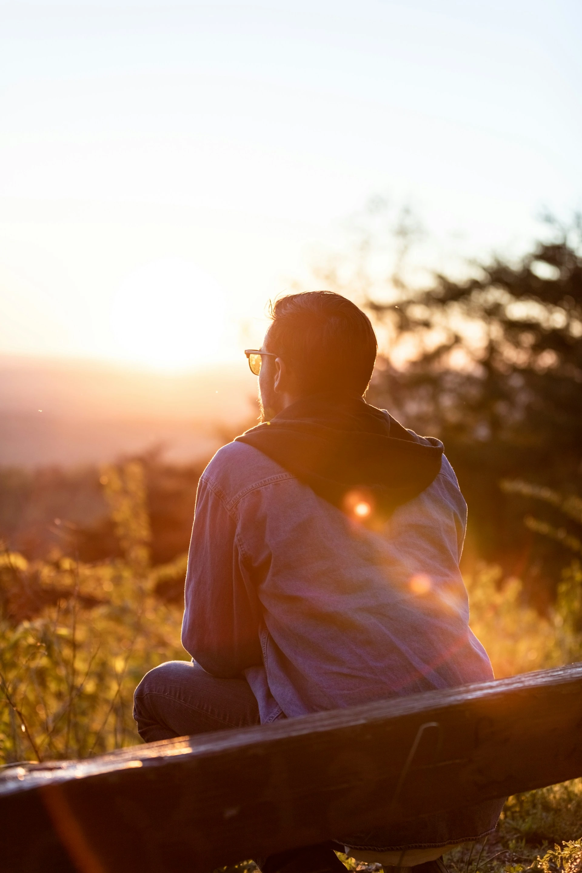 Person wrapped in blanket sitting cross-legged outdoors at sunset, silhouetted against golden light and distant trees.