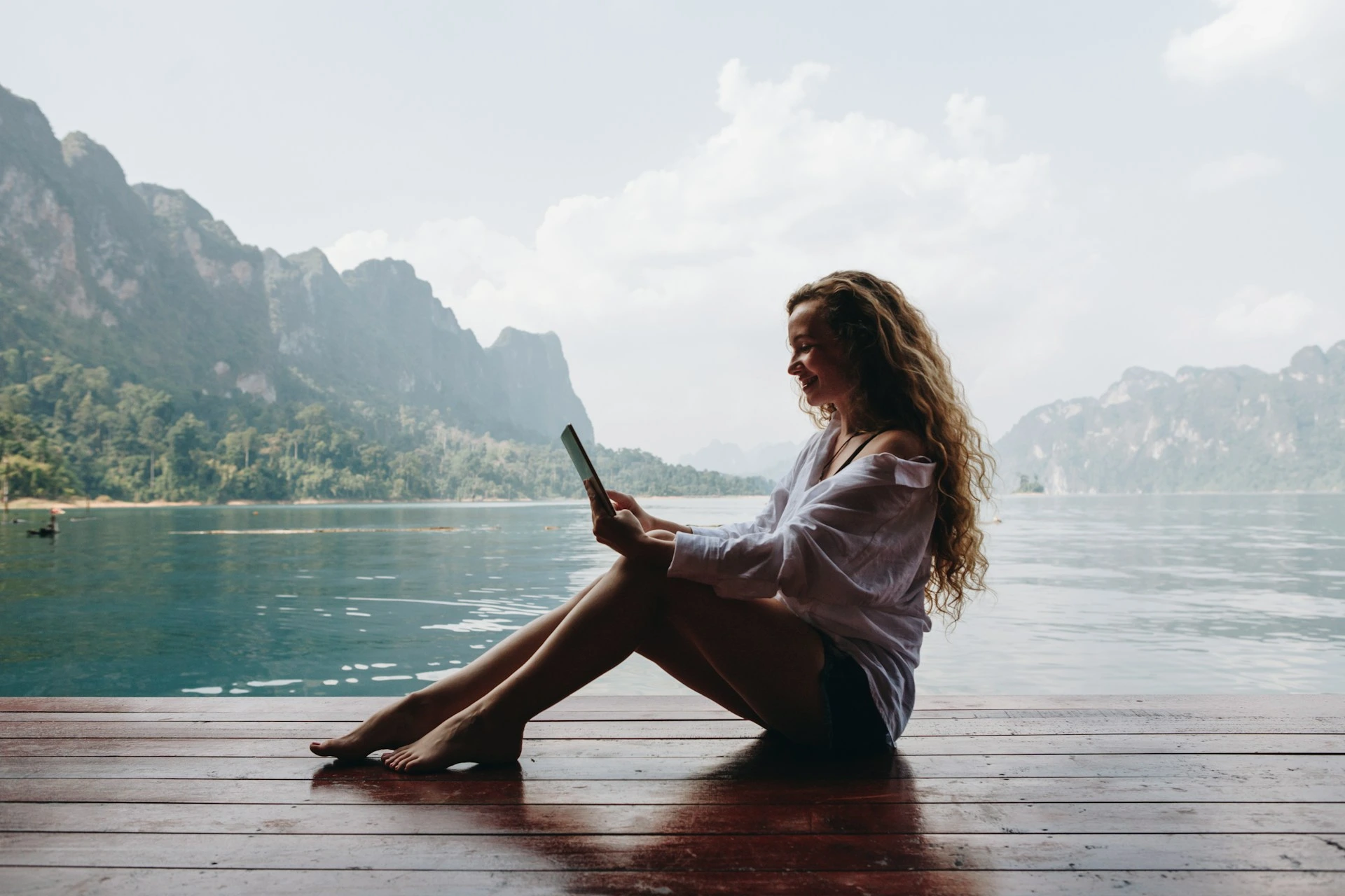 Woman with long curly hair sitting on wooden dock by mountain lake, looking at tablet device in relaxed pose.