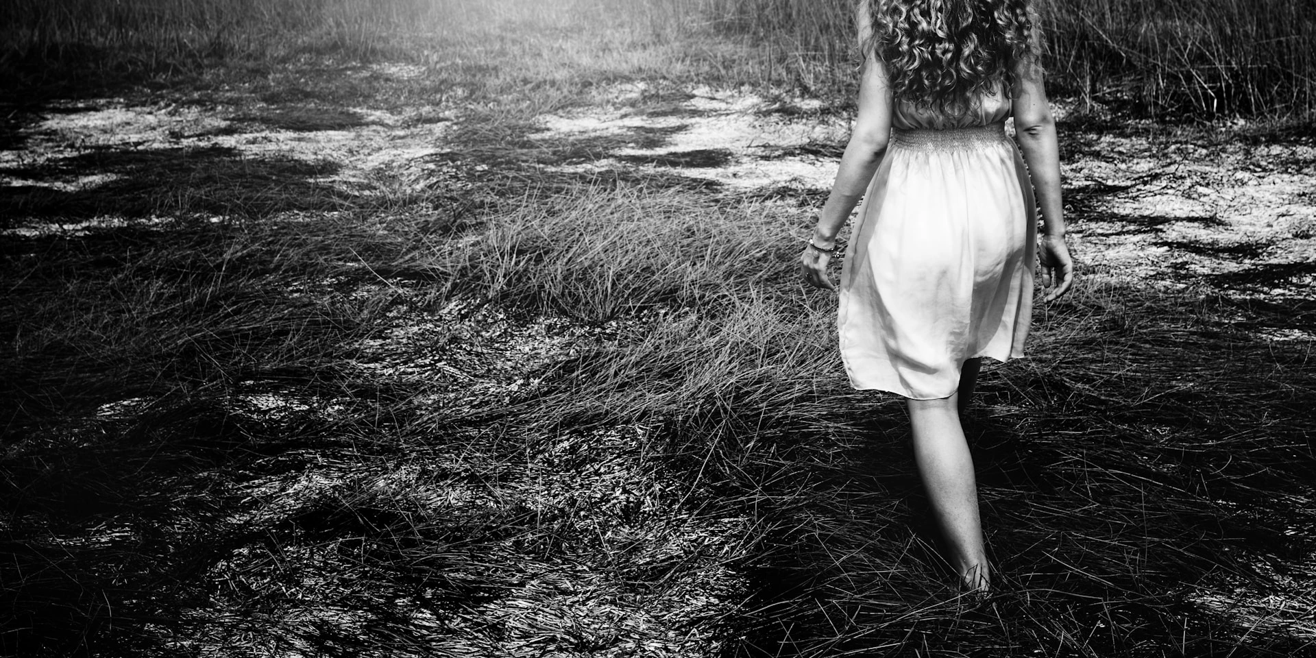Black and white photo of child in white dress walking alone through grassy wooded area, shot from behind.