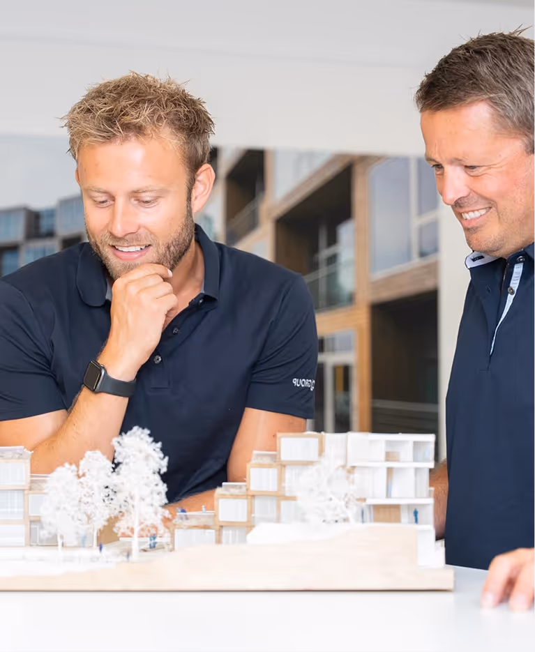 Two men in navy shirts smiling and examining an architectural model of a building with trees on a table.